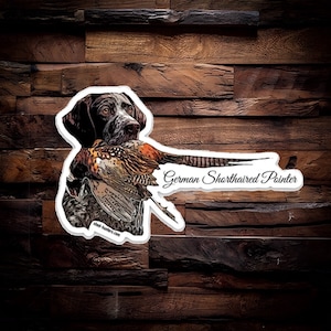 May include: A sticker featuring a German Shorthaired Pointer dog with a pheasant in its mouth. The dog is depicted in a realistic style with brown and white fur. The sticker has a white background with the text "German Shorthaired Pointer" and the website "shef-hunters.com".