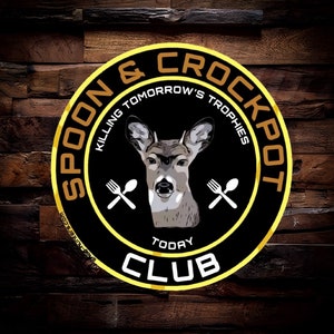 May include: A black and gold circular logo with the text "Spoon & Crockpot" in a gold outline. The logo also includes the text "Killing Tomorrow's Trophies" and "Today's Club" in a white outline. A deer head is in the center of the logo with two crossed utensils.