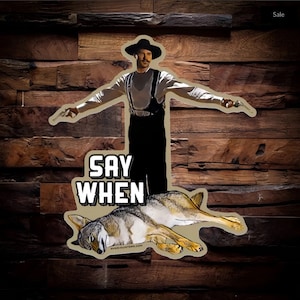 May include: A sticker featuring a man in a cowboy hat and black clothing standing over a wolf with the text "SAY WHEN" on a wooden background.
