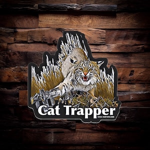 May include: A black and white sticker with a cartoon image of a bobcat with its mouth open and claws out. The text "Cat Trapper" is written in white letters below the image. The text "shed-hunters.com" is written in smaller white letters below the text "Cat Trapper".