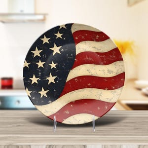 May include: A ceramic plate with a distressed American flag design. The plate is white with red and blue stripes and stars. The plate is on a wooden stand.