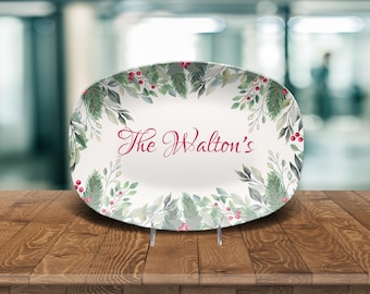 May include: Oval serving platter with a festive design. The white platter features a border of green pine needles, red berries, and gray leaves. The words "The Walton's" are written in red script in the centre.