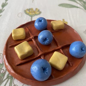 May include: A round brown clay board with a grid pattern for a game, featuring four blue spherical pieces and three yellow square pieces arranged in the squares. The board has a smooth, hand-molded texture with raised lines forming the grid.
