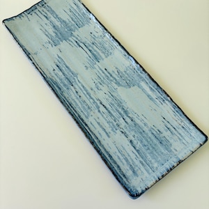 May include: A rectangular ceramic tray featuring a light blue and white glaze with a distressed, brush-stroke texture. The edges of the tray are dark and uneven, highlighting the handcrafted nature of the piece with a smooth, glossy finish.