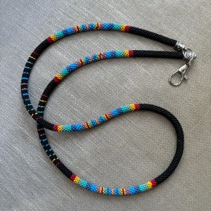 Op de afbeelding: Een zwarte kralenkoord met een kleurrijk kralenpatroon en een zilveren sluiting. Het koord is ongeveer 76 cm lang.