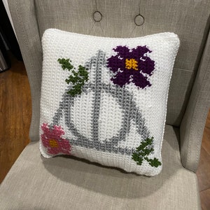 May include: A white crocheted pillow with a gray Deathly Hallows symbol and colorful flowers. The pillow is on a light gray chair.