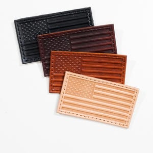 May include: Four rectangular leather patches featuring an embossed American flag design. The patches are arranged in a cascading stack, displaying a gradient of colors from black to dark brown, medium brown, and a light natural tan leather finish.