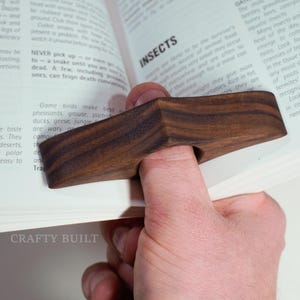 May include: A wooden thumb page holder made of dark brown wood. The holder is shaped like a teardrop and has a hole in the center for the thumb. The holder is resting on a book with the words "BIRDS" and "INSECTS" visible.