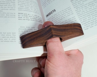 May include: A wooden thumb page holder made of dark brown wood. The holder is shaped like a teardrop and has a hole in the center for the thumb. The holder is resting on a book with the words "BIRDS" and "INSECTS" visible.