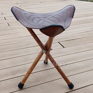 May include: A brown leather folding stool with a Celtic knot design on the seat. The stool has three wooden legs and black rubber feet.