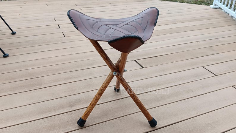 May include: A brown leather folding stool with a Celtic knot design on the seat. The stool has three wooden legs and black rubber feet.
