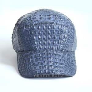 May include: A blue baseball cap made of crocodile leather. The cap has a textured surface and a curved brim. The text "CRAFTY BUILT" is printed on the bottom of the cap.