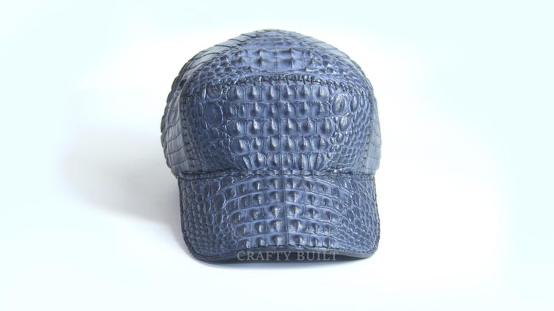 May include: A blue baseball cap made of crocodile leather. The cap has a textured surface and a curved brim. The text "CRAFTY BUILT" is printed on the bottom of the cap.