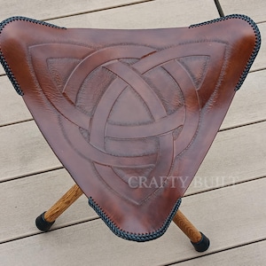 May include: A brown leather folding stool with a Celtic knot design. The stool has three legs and is made of wood. The stool is sitting on a wooden deck.