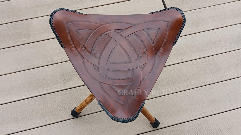 May include: A brown leather folding stool with a Celtic knot design. The stool has three legs and is made of wood. The stool is sitting on a wooden deck.
