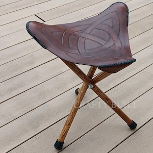 May include: A brown leather folding stool with a Celtic knot design. The stool has three wooden legs and black metal tips.