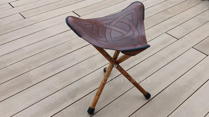 May include: A brown leather folding stool with a Celtic knot design. The stool has three wooden legs and black metal tips.