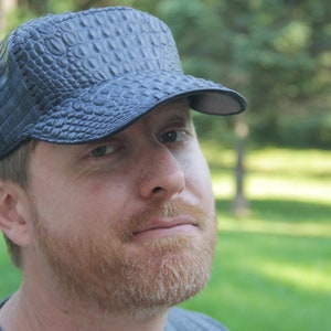 May include: A man wearing a black leather cap with a crocodile skin pattern. The cap has a curved brim and a strap in the back.