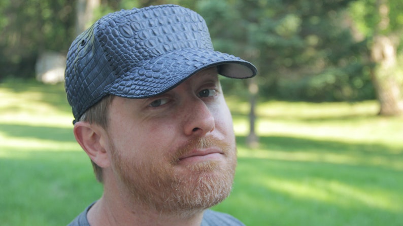 May include: A man wearing a black leather cap with a crocodile skin pattern. The cap has a curved brim and a strap in the back.
