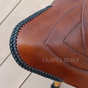 May include: Close-up of a brown leather seat with a black braided edge. The leather has a Celtic knot design and the text "CRAFTY BUILT" is visible.