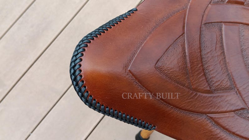 May include: Close-up of a brown leather seat with a black braided edge. The leather has a Celtic knot design and the text "CRAFTY BUILT" is visible.