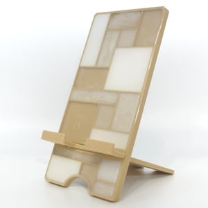 May include: A beige and white geometric pattern phone stand. The stand is angled with a support leg and a ledge to hold a phone. The design features a mosaic of rectangular and square shapes in varying shades of beige and white.