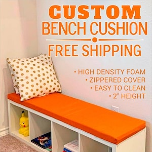 May include: A bright orange rectangular bench cushion with a 2-inch height, featuring a zippered cover and high-density foam. The cushion sits on a white cubby storage unit, accompanied by a white throw pillow with gold polka dots.