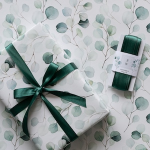 Op de afbeelding: Een ingepakt cadeau met een donkergroen satijnen lint en een bijpassende rol lint. Het cadeaupapier en het lint hebben een aquarel eucalyptusblad ontwerp in groen- en grijstinten op een witte achtergrond.