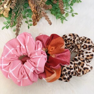 May include: Three fabric scrunchies in pink, red, and leopard print patterns. The pink scrunchie has a white grid design. The red scrunchie has a plaid pattern. The leopard print scrunchie has a brown and black spotted design.