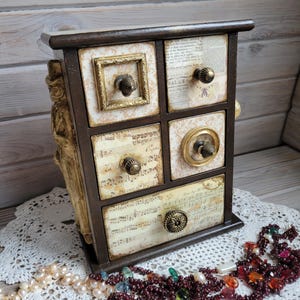May include: A small, dark brown wooden chest of drawers with six drawers. Each drawer has a unique design, including patterned paper, musical notes, and decorative knobs. A gold-colored statue is on the left side. A string of pearls and a beaded necklace are in the foreground.