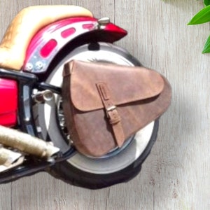 Puede incluir: Una alforja de cuero marrón con cierre de hebilla está montada en la rueda trasera de una motocicleta roja y negra. La motocicleta tiene un asiento beige y tubos de escape cromados. El fondo es una veta de madera clara.
