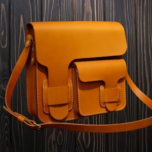 May include: A brown leather satchel bag with a flap closure and two front pockets. The bag has a long adjustable strap.