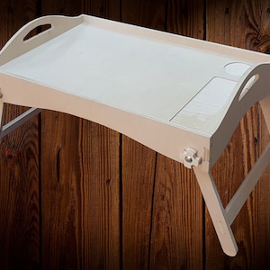 May include: White wooden tray table with a raised edge and two side compartments. The table has foldable legs and a decorative design on the corners.