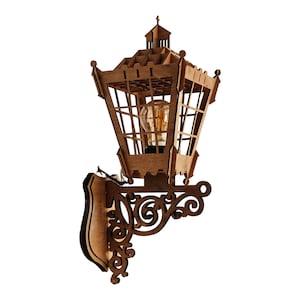 Puede incluir: Una lámpara de pared de madera con un diseño decorativo y ornamentado. La lámpara tiene una estructura en forma de jaula alrededor de la bombilla y un soporte decorativo con volutas.