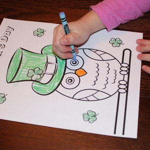 May include: A child's hand coloring a St. Patrick's Day coloring page with a blue crayon. The page features an owl wearing a green hat with shamrocks and the words "St. Patrick's Day".