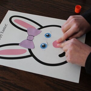 May include: A white paper craft with a black outline of a bunny rabbit. The bunny has pink ears, a purple bow, blue eyes, and a pink nose. The paper is on a wooden table. The text "Happy Easter!" is written on the paper.