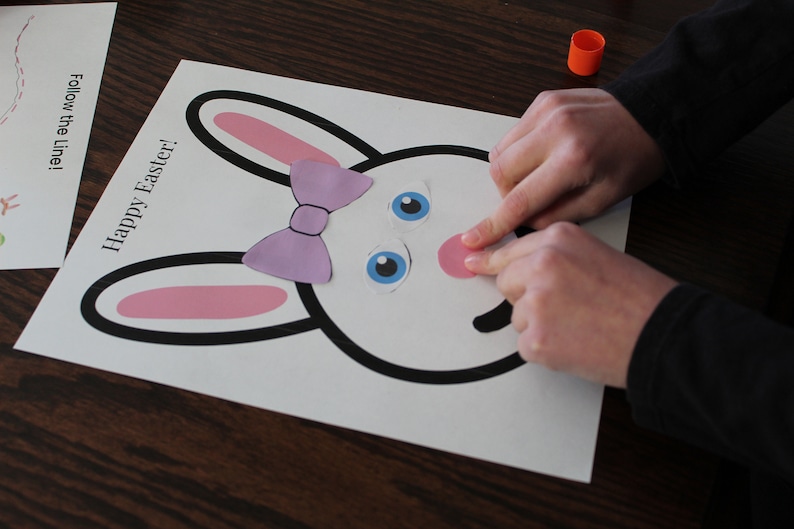 May include: A white paper craft with a black outline of a bunny rabbit. The bunny has pink ears, a purple bow, blue eyes, and a pink nose. The paper is on a wooden table. The text "Happy Easter!" is written on the paper.