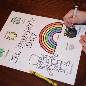 May include: A St. Patrick's Day coloring page with a rainbow, leprechaun, shamrock, horseshoe, and pot of gold. The words "St. Patrick's Day" are printed on the page. A person is coloring the pot of gold with a black crayon.