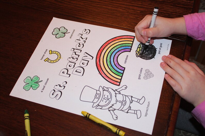 May include: A St. Patrick's Day coloring page with a rainbow, leprechaun, shamrock, horseshoe, and pot of gold. The words "St. Patrick's Day" are printed on the page. A person is coloring the pot of gold with a black crayon.