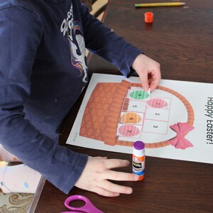 May include: A child is gluing colourful Easter eggs to a paper basket with the words "Happy Easter!" printed on it. The basket has a pink bow and the eggs are numbered 1 through 7.