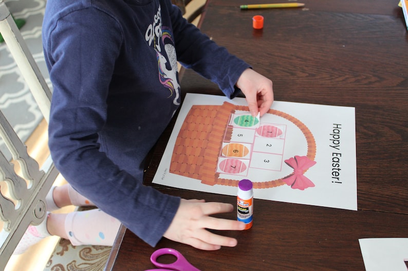 May include: A child is gluing colourful Easter eggs to a paper basket with the words "Happy Easter!" printed on it. The basket has a pink bow and the eggs are numbered 1 through 7.