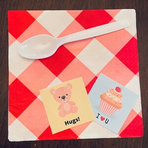 May include: Two small cards with cute illustrations on a red and white checkered napkin. One card features a pink teddy bear with the text "Hugs!" and the other card features a pink cupcake with a cherry on top and the text "I &hearts; U".