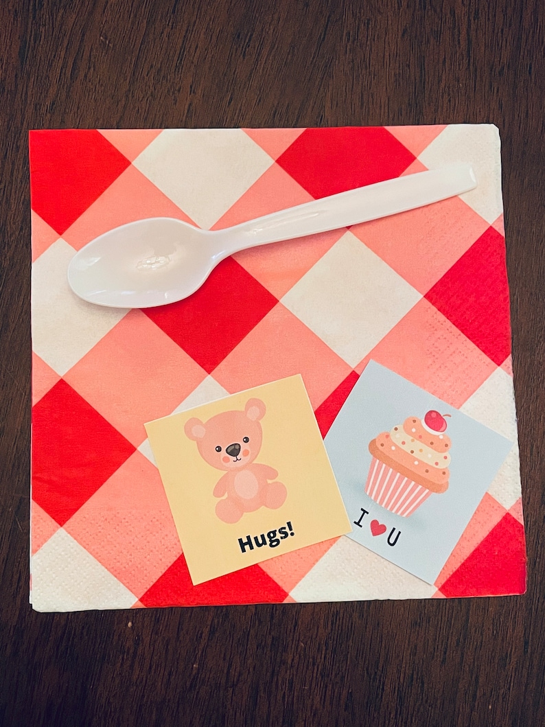 May include: Two small cards with cute illustrations on a red and white checkered napkin. One card features a pink teddy bear with the text "Hugs!" and the other card features a pink cupcake with a cherry on top and the text "I &hearts; U".