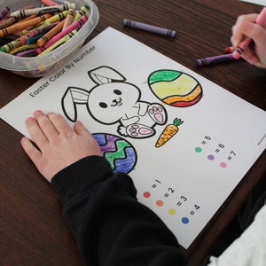 May include: A child is colouring in a printable Easter colouring page with crayons. The page features a bunny rabbit, two eggs, and a carrot. The page has a colour key with numbers and corresponding colours.