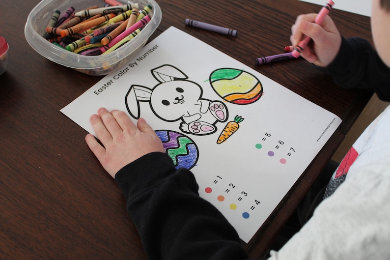 May include: A child is colouring in a printable Easter colouring page with crayons. The page features a bunny rabbit, two eggs, and a carrot. The page has a colour key with numbers and corresponding colours.