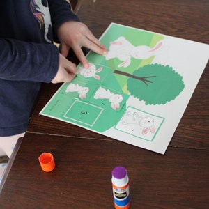 May include: A child is putting together a puzzle of a white bunny hopping over a green tree. The puzzle pieces are numbered 1, 2, 3, and 4. The child is using glue to secure the pieces.