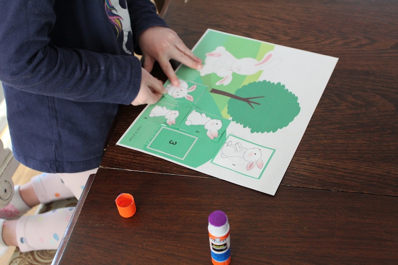 May include: A child is putting together a puzzle of a white bunny hopping over a green tree. The puzzle pieces are numbered 1, 2, 3, and 4. The child is using glue to secure the pieces.