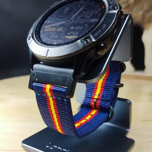 Op de afbeelding: Een zwart en zilveren smartwatch met een blauw, geel en rood gestreept nylon horlogebandje. Het horloge display toont de tijd, datum en andere informatie. Het horloge staat op een grijze standaard.