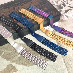 May include: Seven different metal watch bands in various colors, including silver, gold, black, blue, and iridescent. The bands are laid out on a wooden surface with a vintage design.