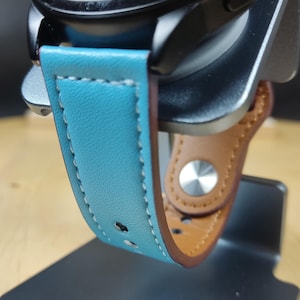 May include: A black smartwatch with a blue leather band and a brown leather strap. The watch is on a gray metal stand with the word "remarkable" engraved on it.
